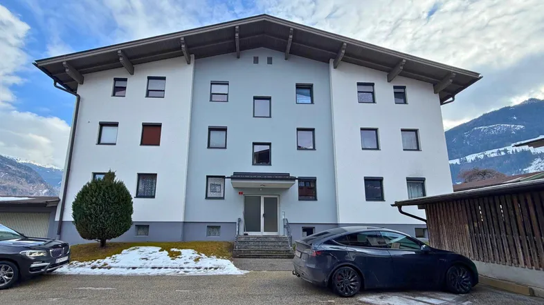 Modernes Mehrfamilienhaus mit heller Fassade und Satteldach, umgeben von winterlicher Berglandschaft.