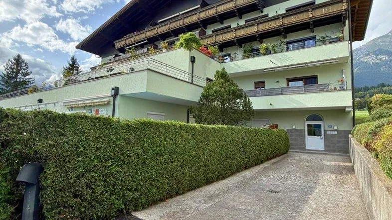 Mehrstöckiges Wohngebäude mit Balkonen und gepflegter Hecke, umgeben von grüner Landschaft und Bergen.