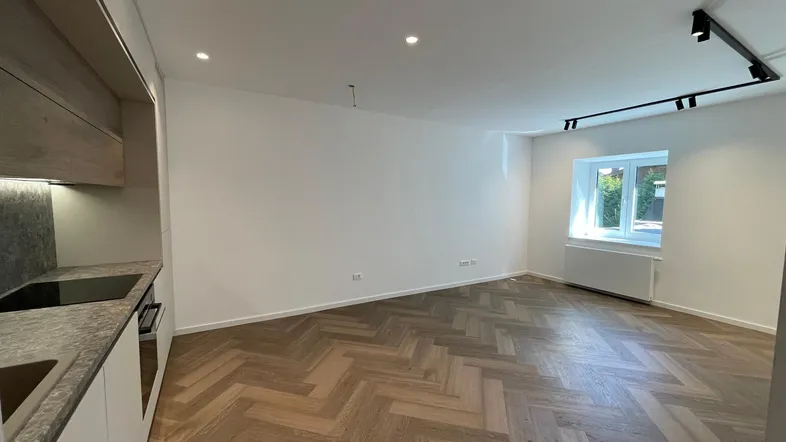 Heller Wohnbereich mit integrierter Küche, Fischgrätparkett und Fenster mit Grünblick.