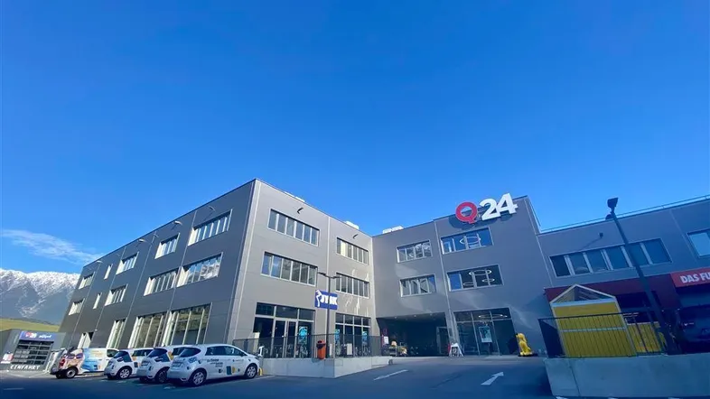 Modernes Geschäftsgebäude mit Parkplätzen davor und Bergen im Hintergrund unter blauem Himmel.