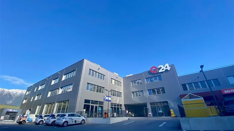 Modernes Geschäftsgebäude mit großzügigen Fensterfronten, Parkplätzen und beeindruckendem Bergblick unter blauem Himmel.
