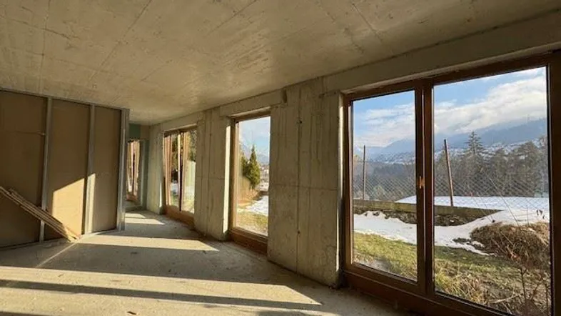 Unfertiger Innenraum mit Betonwänden und großen Fenstern, die einen Blick auf die verschneite Landschaft bieten.