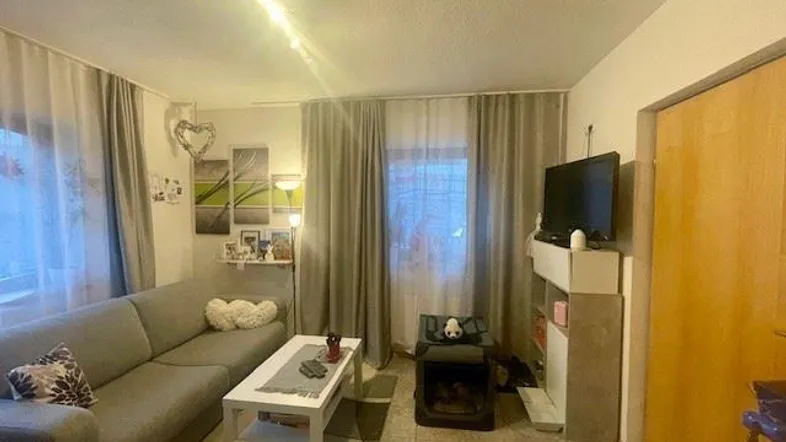 Gemütliches Wohnzimmer mit Sofa, Couchtisch und Fernseher, ausgestattet mit Fliesenboden und Vorhängen.