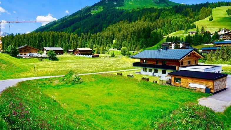 Haus in alpiner Landschaft mit grünen Wiesen und bewaldeten Bergen im Hintergrund.