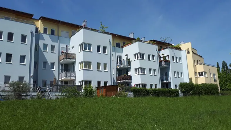 Modernes Mehrfamilienhaus mit hellblauen und gelben Fassaden, umgeben von einer grünen Wiese unter blauem Himmel.