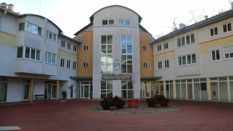 Ansicht des Gemeindeamtes mit gepflastertem Vorplatz und modernen Gebäudeflügeln unter bewölktem Himmel.