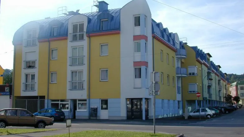 Mehrstöckiges Gebäude mit gelber Fassade, blauen Akzenten und Mansarddach in städtischer Umgebung.