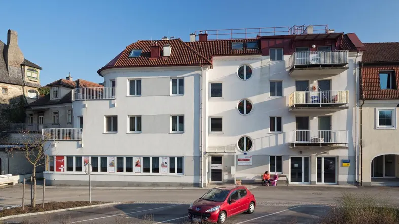 Mehrstöckiges Gebäude mit weißer Fassade, roten Dachziegeln und mehreren Balkonen, davor ein Parkplatz.