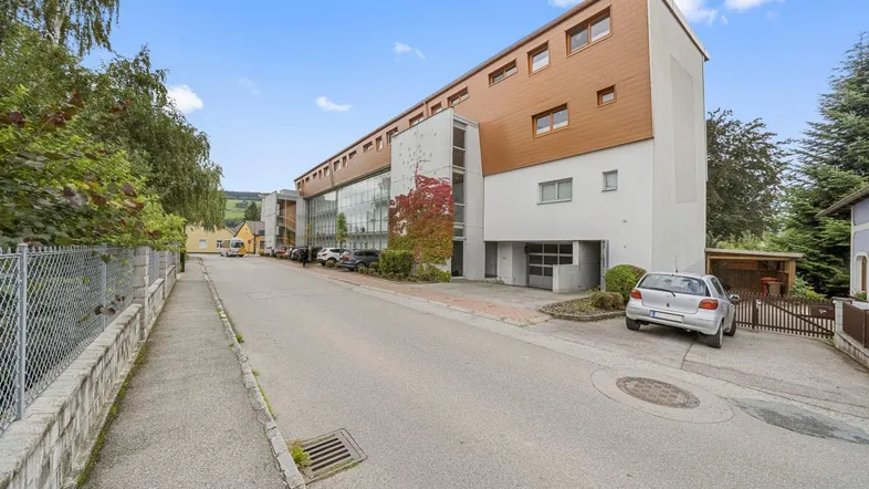 Modernes Gebäude mit langer Fassade, Glasfronten und Holzverkleidung, umgeben von einer Straße und Parkplätzen.