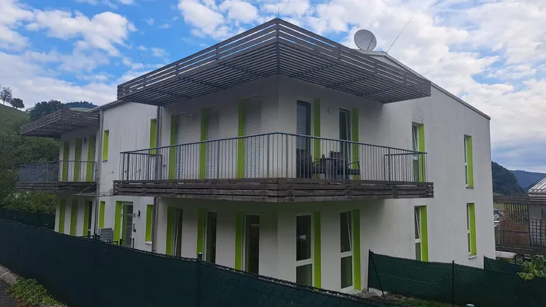 Modernes Wohngebäude mit weißer Fassade, grünen Akzenten und mehreren Balkonen vor hügeliger Landschaft.