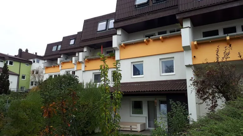 Mehrfamilienhaus mit braunem Mansarddach, gelben Balkonen und grüner Gartenanlage im Vordergrund.