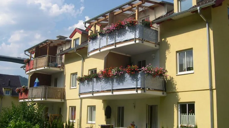 Mehrfamilienhaus mit gelber Fassade, roten Dächern und blumengeschmückten Balkonen in sonniger Lage.