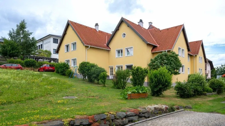 Gelbes Mehrfamilienhaus mit rotem Ziegeldach und gepflegter Grünfläche in ländlicher Umgebung.