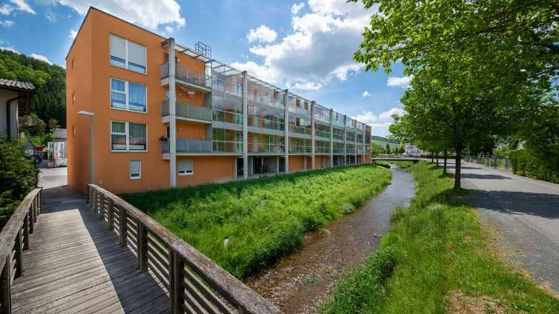 Modernes Wohngebäude mit orangefarbener Fassade, Balkonen und einem Bachlauf im Vordergrund unter blauem Himmel.