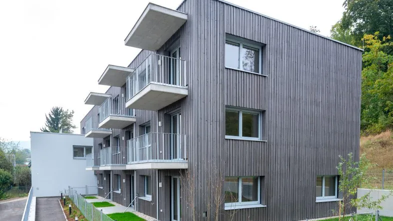 Modernes Mehrfamilienhaus mit Holzfassade und Balkonen, umgeben von Grünflächen und Bäumen.