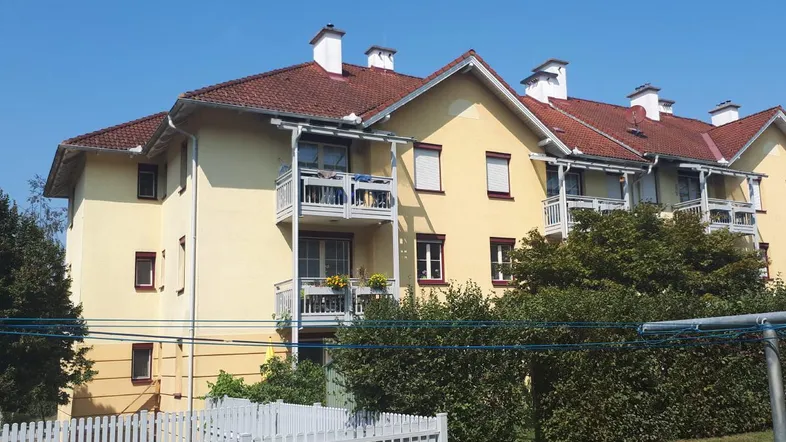Mehrfamilienhaus mit gelber Fassade, roten Ziegeldächern und mehreren Balkonen unter blauem Himmel.