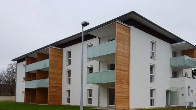 Modernes Mehrfamilienhaus mit weißer Fassade, Holzverkleidung und Balkonen mit Glasbrüstung.