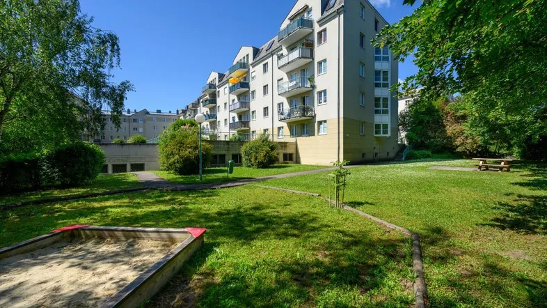 Mehrfamilienhaus mit Balkonen, umgeben von einer grünen Gartenanlage und einem Sandkasten.