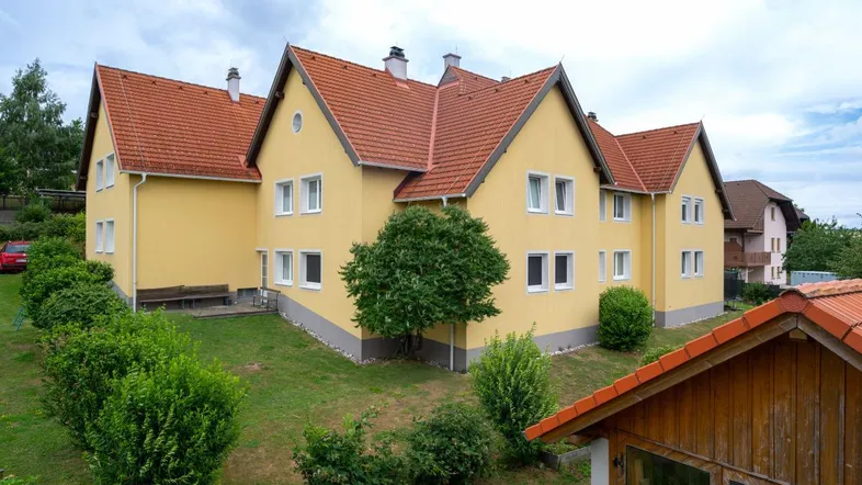 Gelbes Mehrfamilienhaus mit roten Ziegeldächern und gepflegtem Gartenbereich unter bewölktem Himmel.