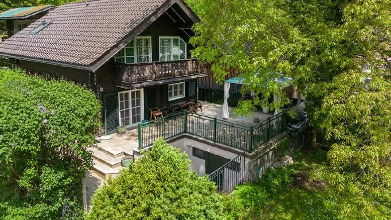 Traditionelles Haus mit großer Terrasse, umgeben von üppigem Grün und Bäumen.