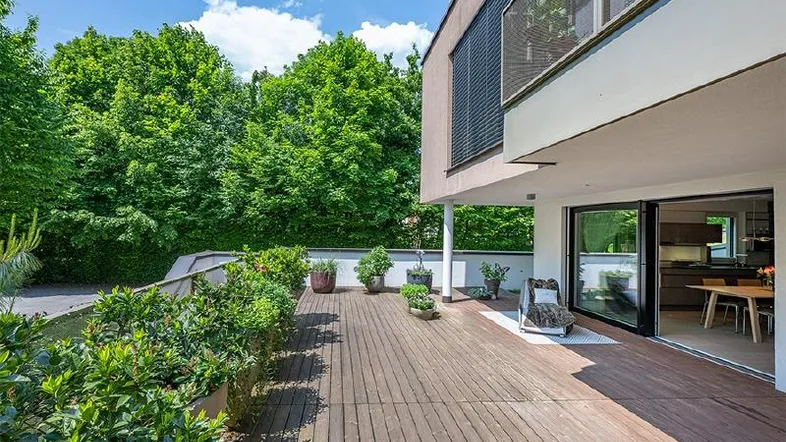 Große Holzterrasse mit Pflanzen und direktem Zugang zum modernen Wohnbereich, umgeben von viel Grün.