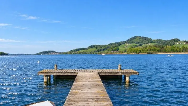 Holzsteg führt in einen ruhigen See mit grünen Hügeln unter blauem Himmel.