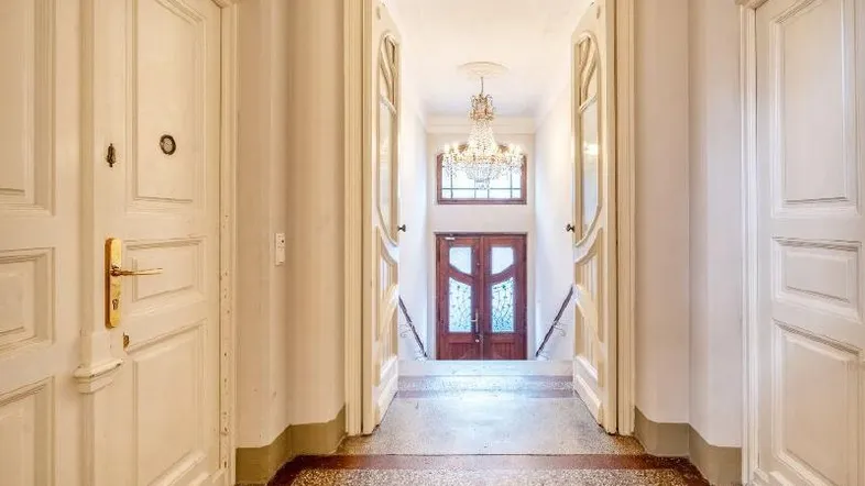Elegante Eingangshalle mit Kronleuchter, hohen Türen und Terrazzoboden, die zu einem weiteren Bereich führt.