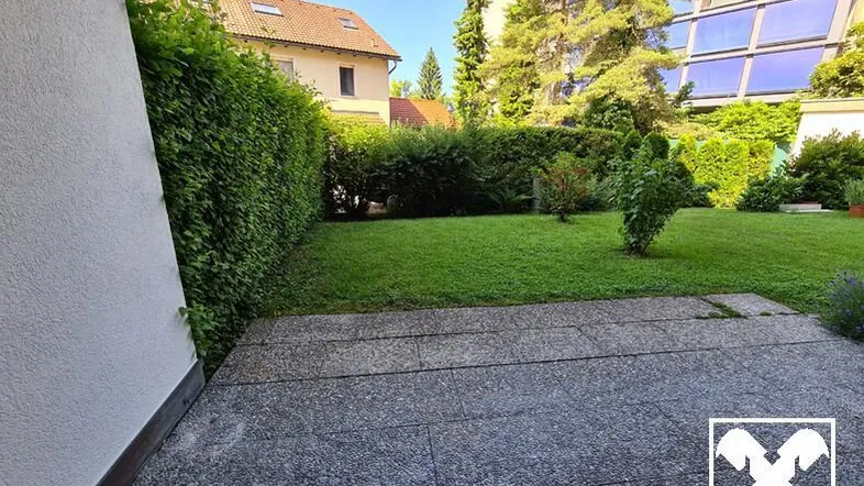 Gepflasterte Terrasse mit Rasenfläche und hoher Hecke, umgeben von viel Grün.