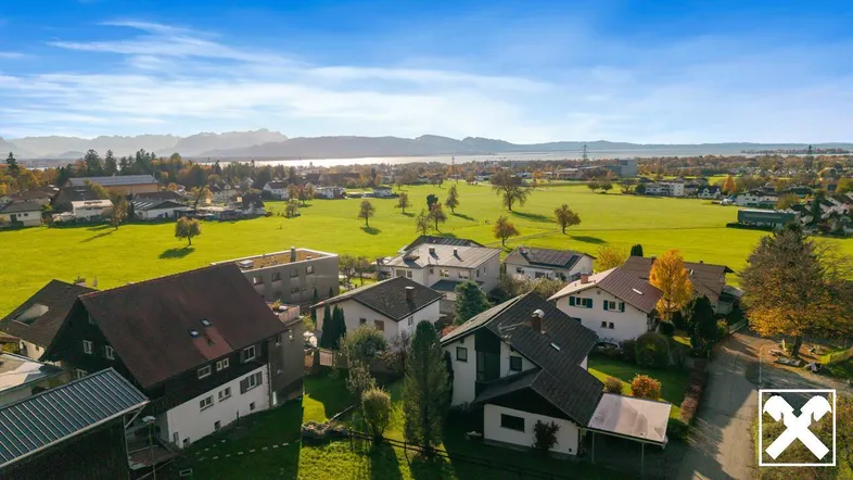 Panoramablick auf eine Wohnsiedlung mit grünen Feldern, See und Bergen im Hintergrund.