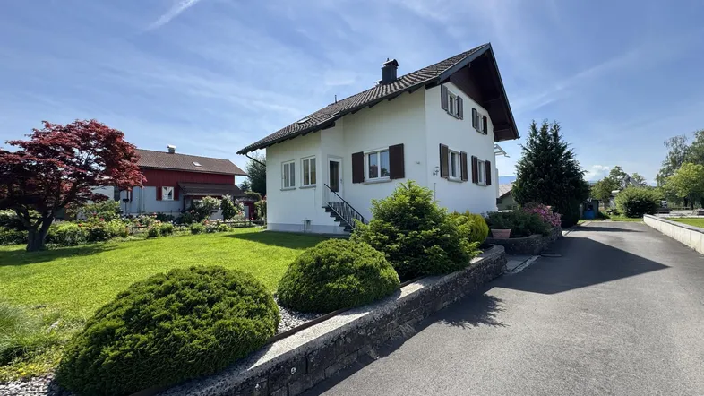 Weißes Einfamilienhaus mit Satteldach, gepflegtem Vorgarten und Zufahrt unter blauem Himmel.