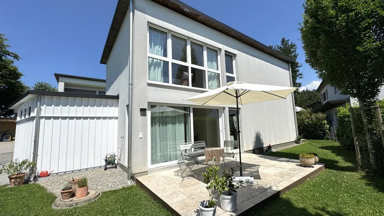 Moderne Hausfassade mit sonniger Terrasse, Gartenmöbeln und gepflegtem Rasenbereich.
