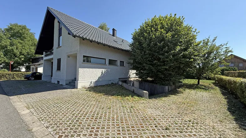 Einfamilienhaus mit Satteldach, gepflasterter Einfahrt und grüner Umgebung unter blauem Himmel.