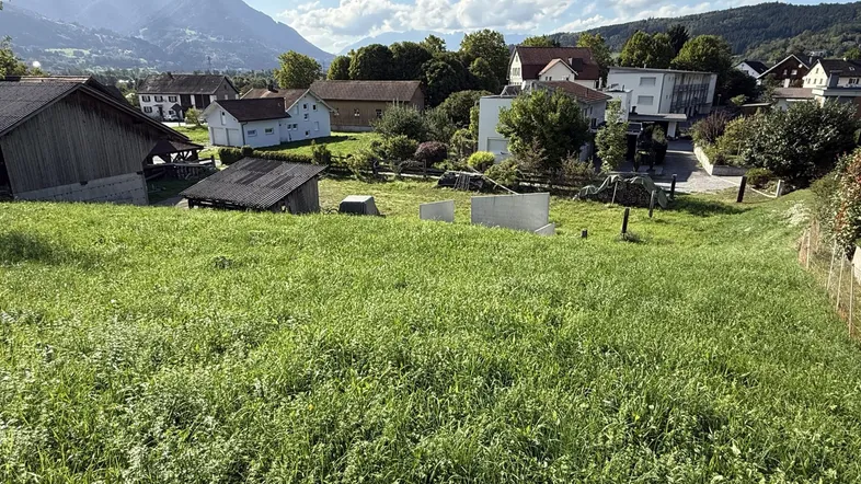 Grünes Grundstück mit Blick auf umliegende Wohnhäuser und Berge unter blauem Himmel.
