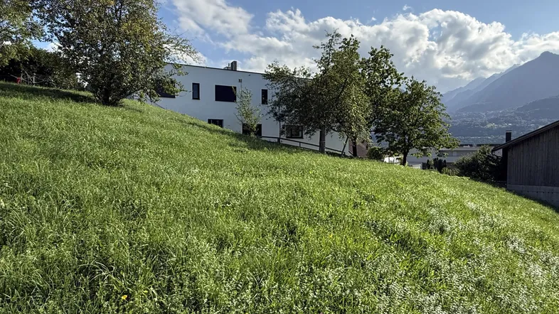 Grüne Wiese mit modernem Gebäude im Hintergrund und Bergblick unter blauem Himmel.