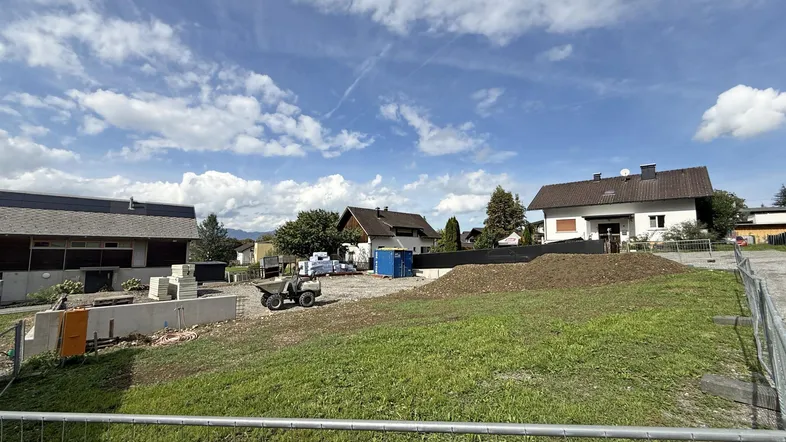 Blick auf ein Baugrundstück mit beginnenden Fundamentarbeiten und umliegenden Häusern unter bewölktem Himmel.