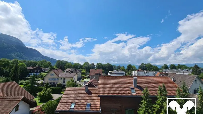 Panoramablick über Dächer, grüne Bäume und Berge unter einem strahlend blauen Himmel.