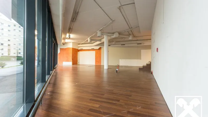 Großer, leerer Gewerberaum mit dunklem Holzboden, hohen Decken und großen Fenstern, die viel Tageslicht hereinlassen.