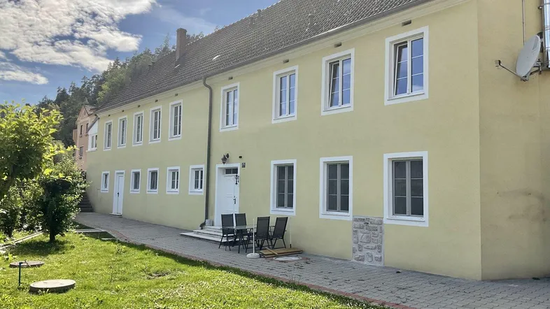 Gelbe Fassade eines Mehrfamilienhauses mit gepflegtem Vorgarten und Sitzgelegenheiten im Freien unter blauem Himmel.