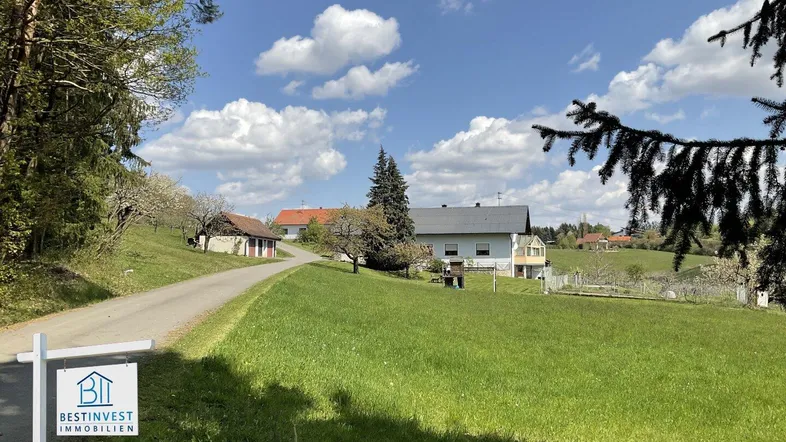 Ländliche Umgebung mit einem Haus, umgeben von grünen Wiesen und Bäumen unter blauem Himmel.