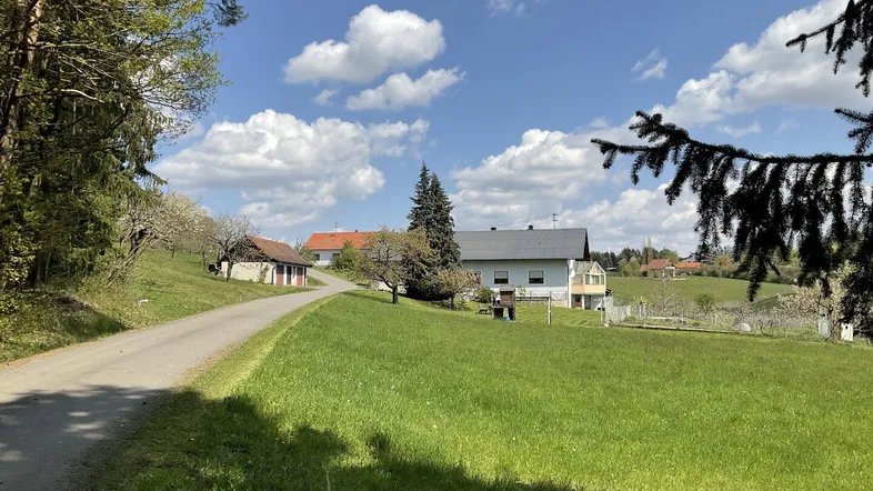 Ländliches Anwesen mit Wohnhaus und Nebengebäuden, umgeben von grünen Wiesen und Bäumen unter blauem Himmel.