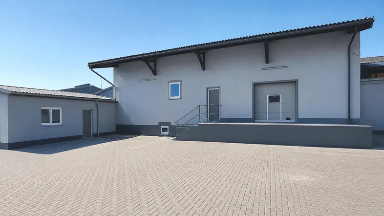Graues Industriegebäude mit Laderampe und großem gepflasterten Vorplatz unter blauem Himmel.