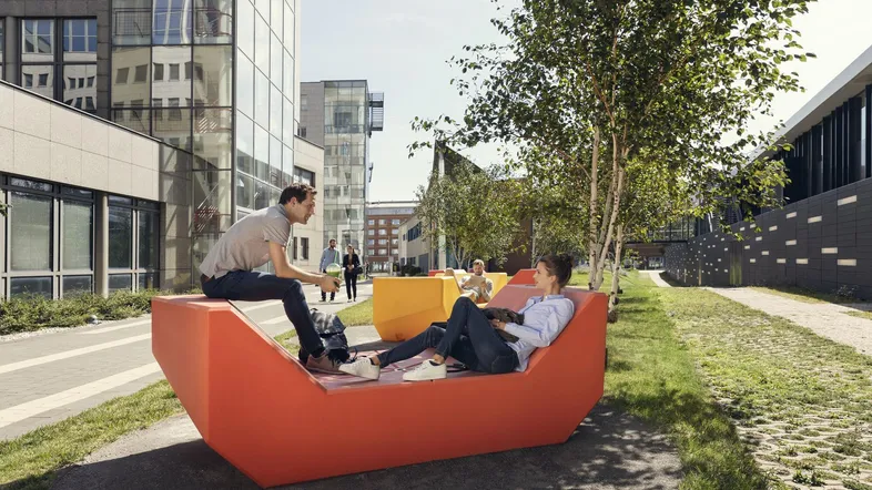 Zwei Personen entspannen auf modernen orangefarbenen Sitzmöbeln in einem begrünten Innenhof mit moderner Architektur im Hintergrund.