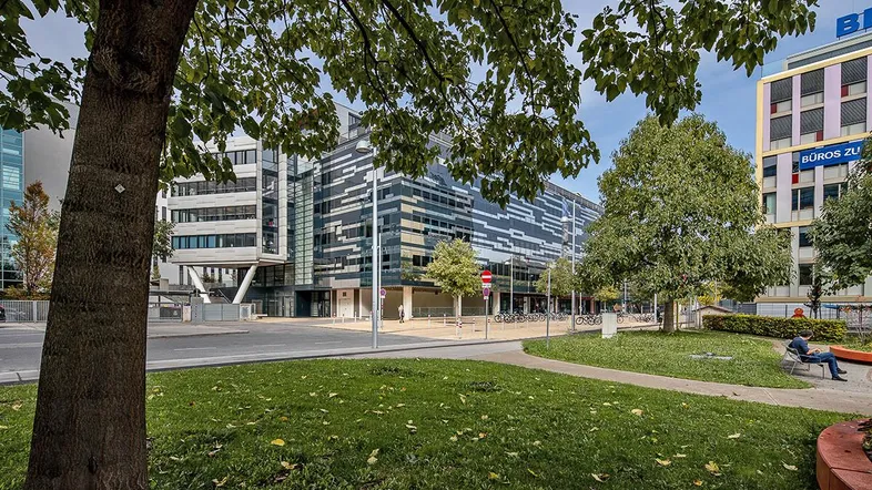 Moderne Bürogebäude mit Glasfassaden und Grünflächen in urbaner Umgebung, umgeben von Bäumen und einem Park.