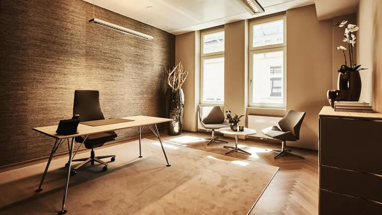 Modernes Büro mit Schreibtisch, bequemen Stühlen und großen Fenstern, die viel Tageslicht hereinlassen.