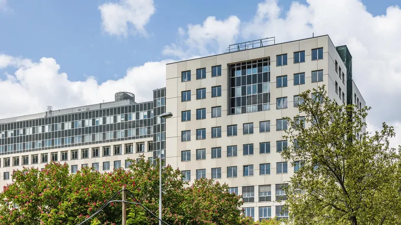Modernes Bürogebäude mit zahlreichen Fenstern und einer hellen Fassade, umgeben von grünen Bäumen unter blauem Himmel.