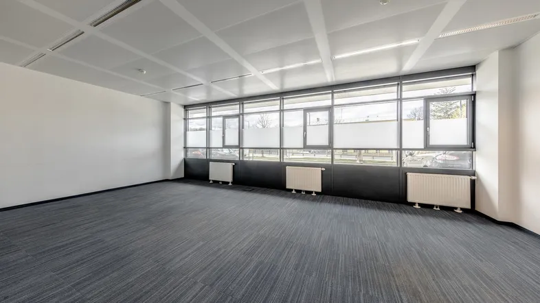 Geräumiges Büro mit Teppichboden, großen Fenstern und Heizkörpern unter der Fensterbank.