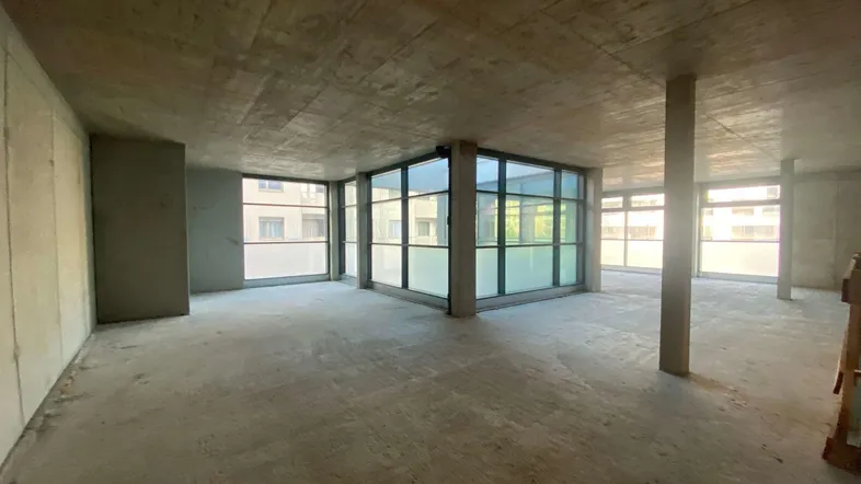 Rohbau-Innenraum mit Sichtbetonwänden und großen Fensterfronten, die viel Tageslicht hereinlassen.
