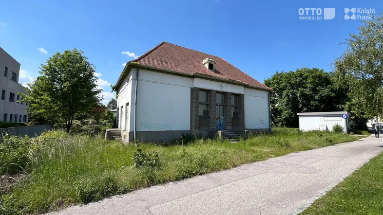 Gebäude mit rotem Ziegeldach und weißer Fassade, umgeben von Gras und Bäumen unter blauem Himmel.