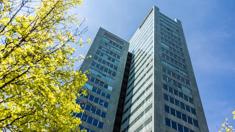 Modernes Hochhaus mit vielen Fenstern und einer Fassade aus Glas und Metall, umgeben von grünen Bäumen unter blauem Himmel.