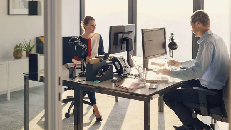 Zwei Personen arbeiten in einem modernen Büro mit großen Fenstern, die viel natürliches Licht hereinlassen.